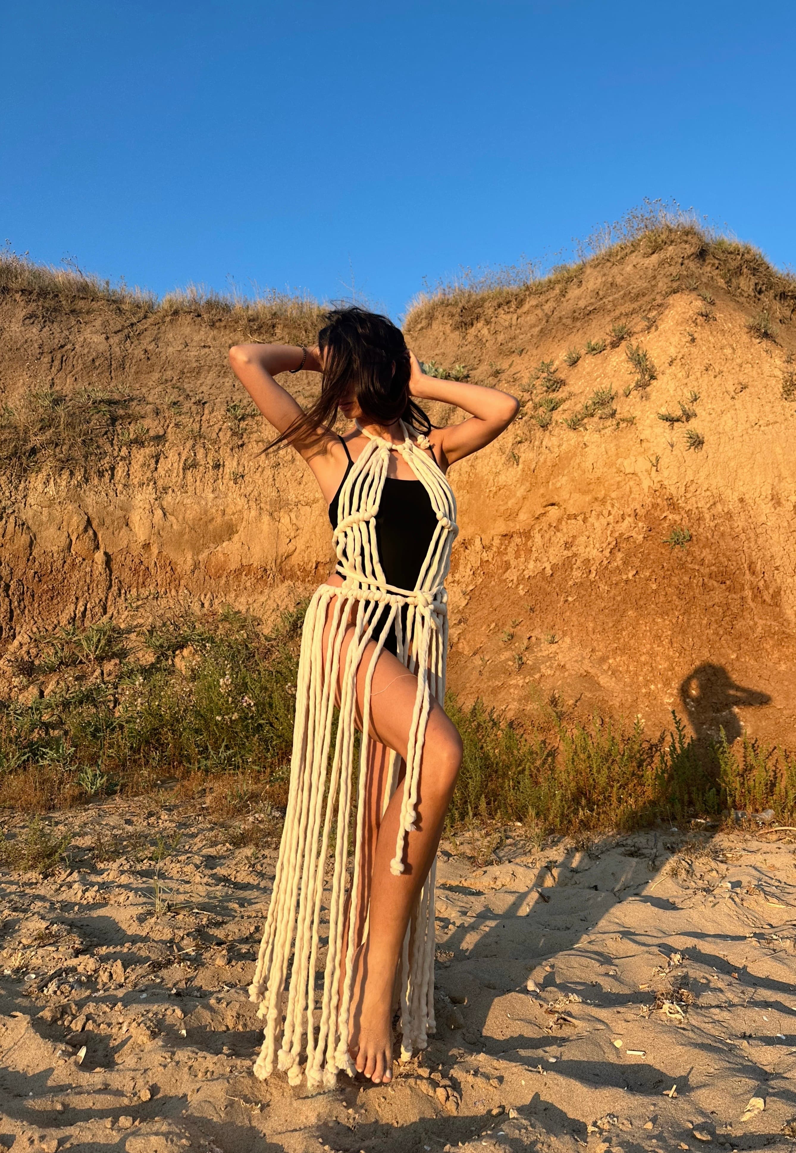 A woman wearing a beige knit fringe beach dress posing outdoors against a sandy cliff.