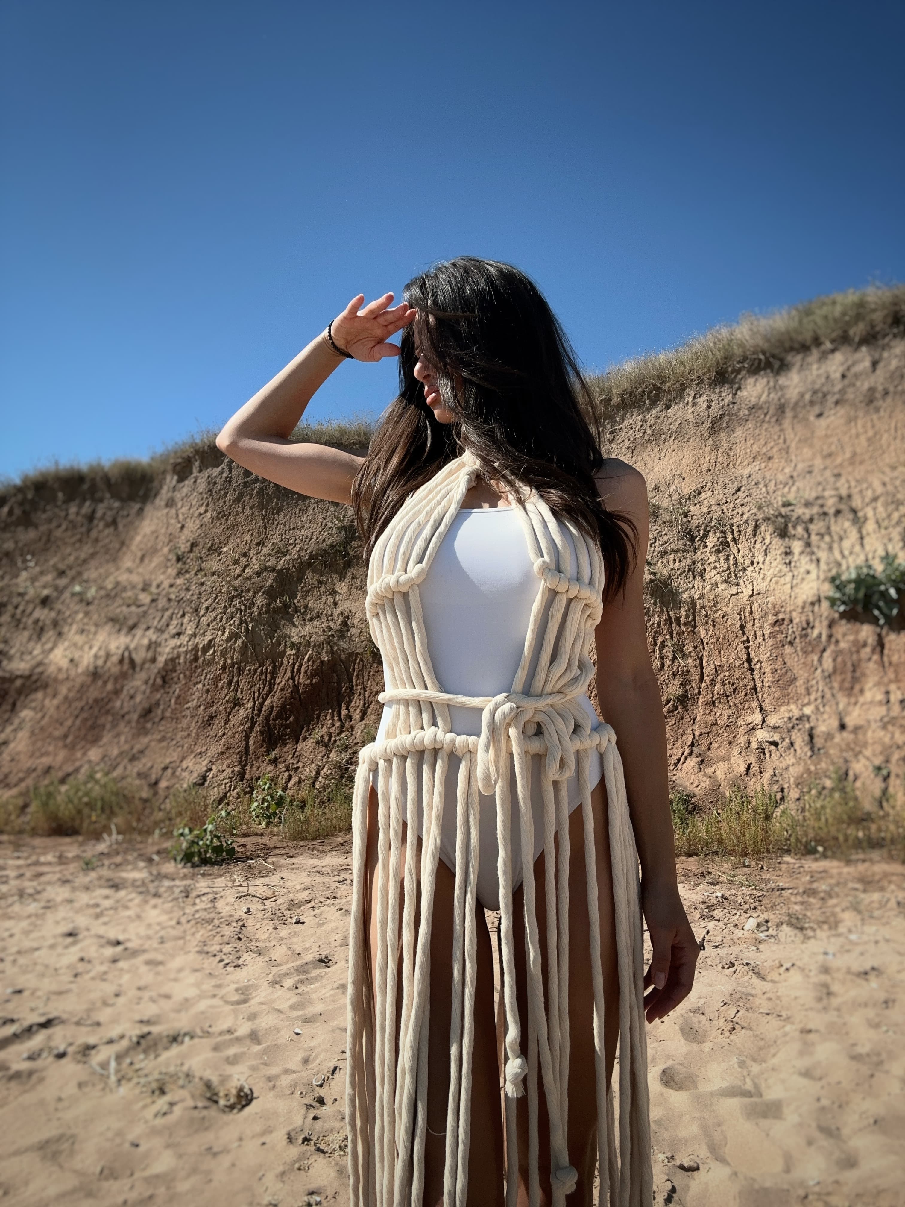 A woman on a sandy beach wears a beige sheer knit fringe long dress over a white swimsuit.