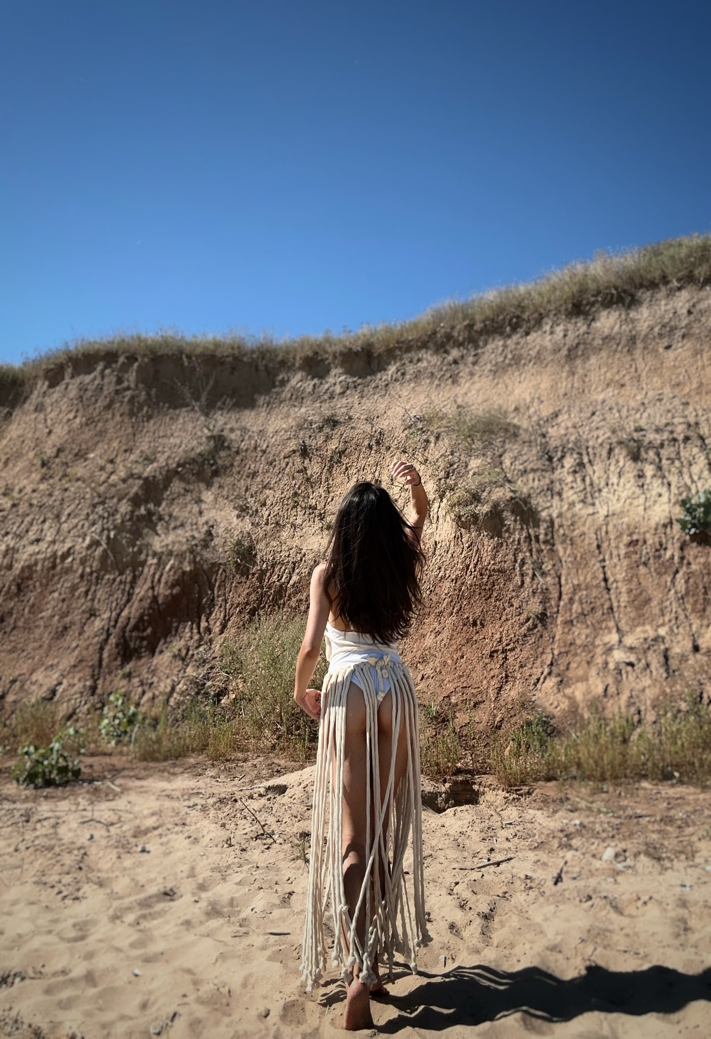A woman stands facing a sand cliff in a cream-colored fringe long knit dress and white bikini bottoms.