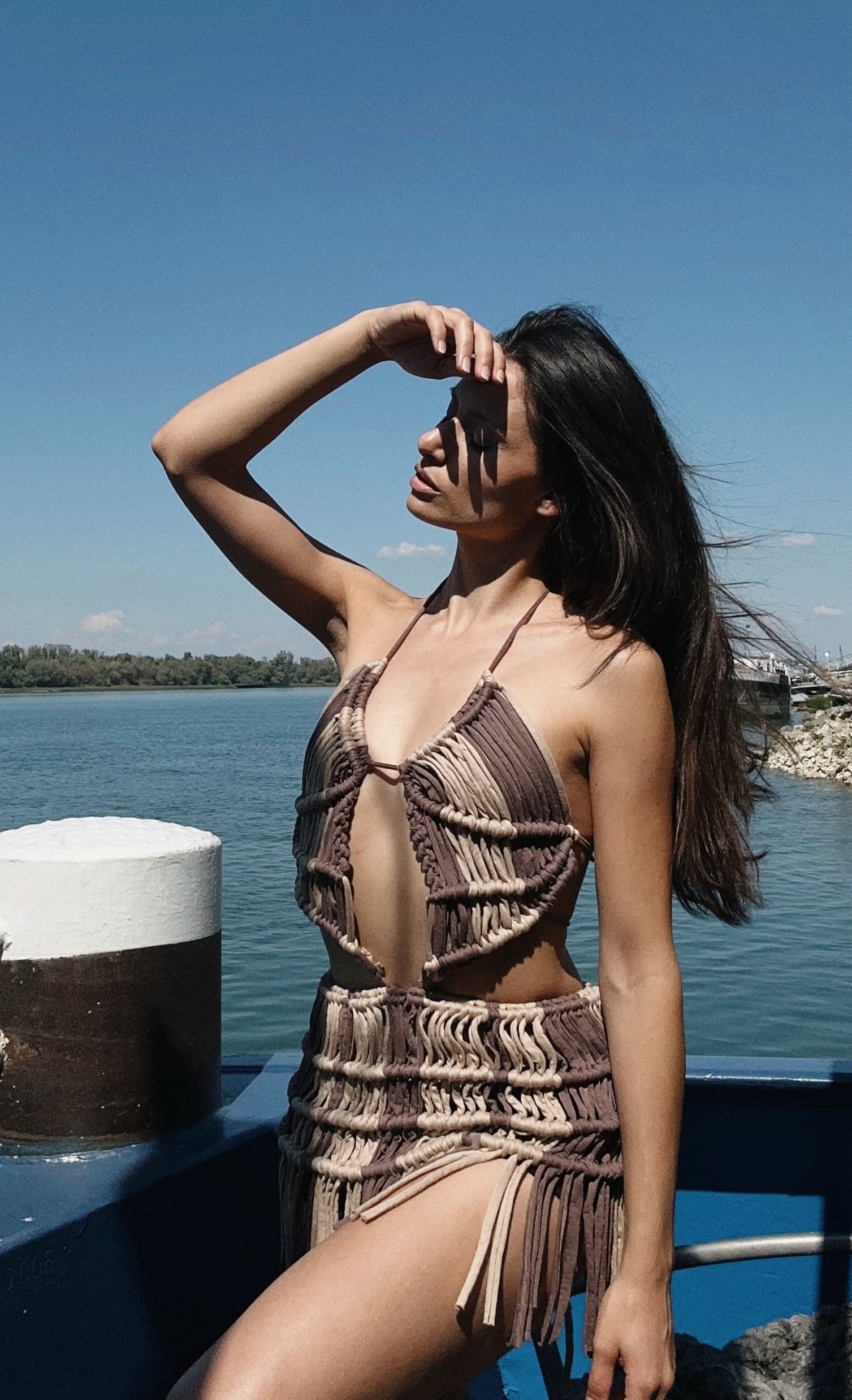 A woman wearing a brown and beige macrame fringe mini dress posing near water.