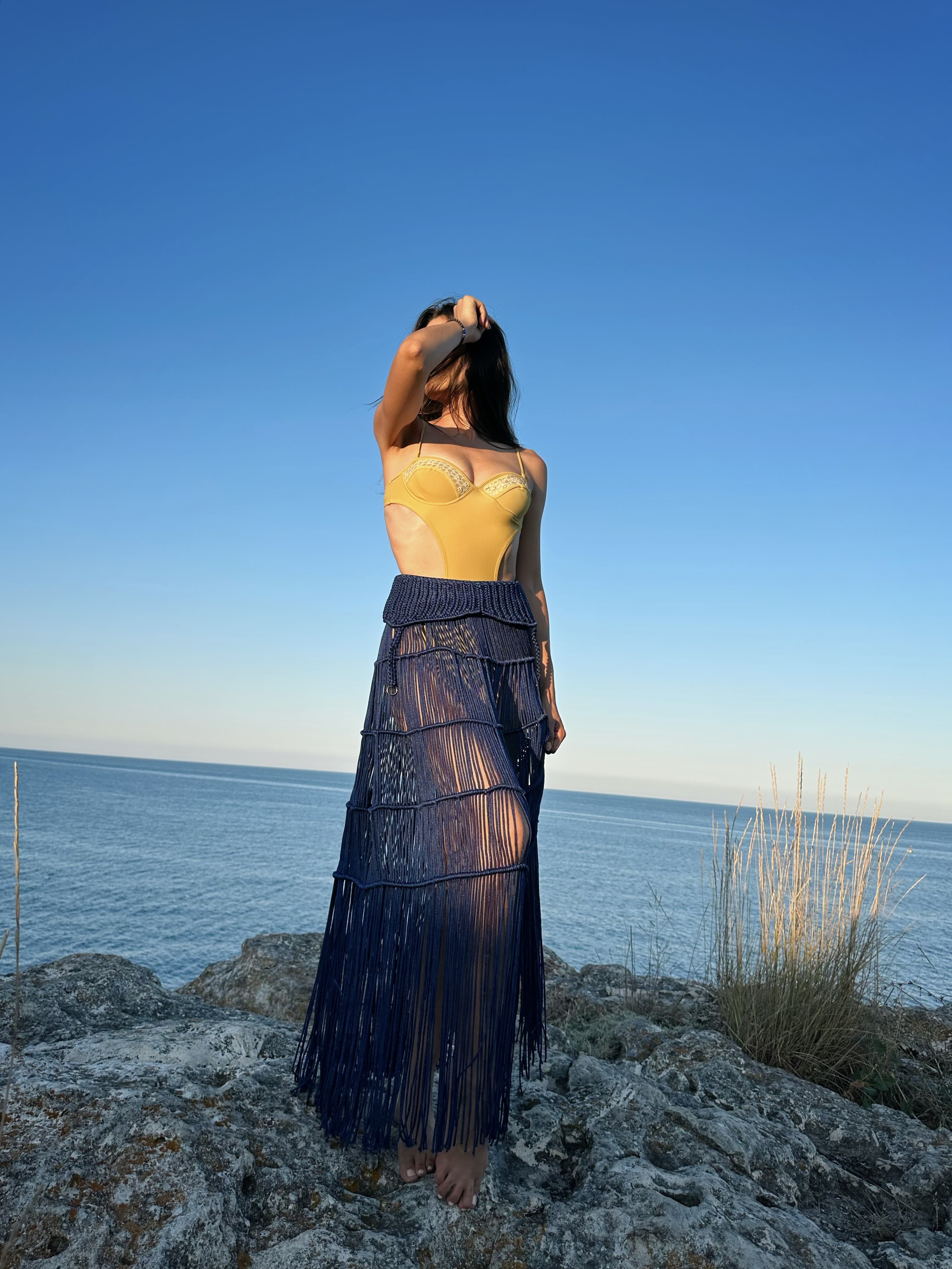 A woman wearing a yellow swimsuit and a long dark blue fringed skirt standing on coastal rocks by the sea.