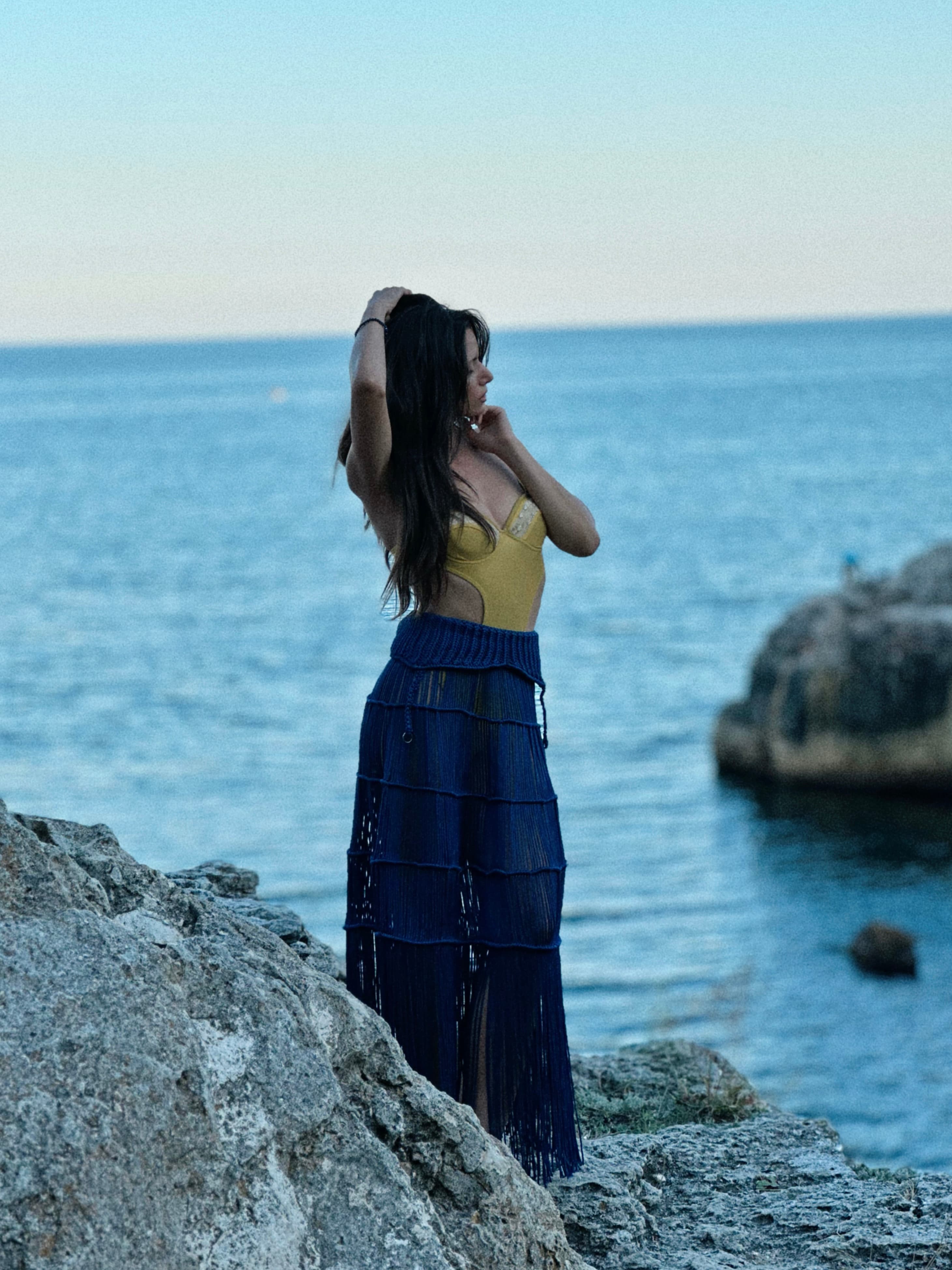 A woman wearing a yellow top and blue knitted beach skirt stands on rocky cliffs by the sea.