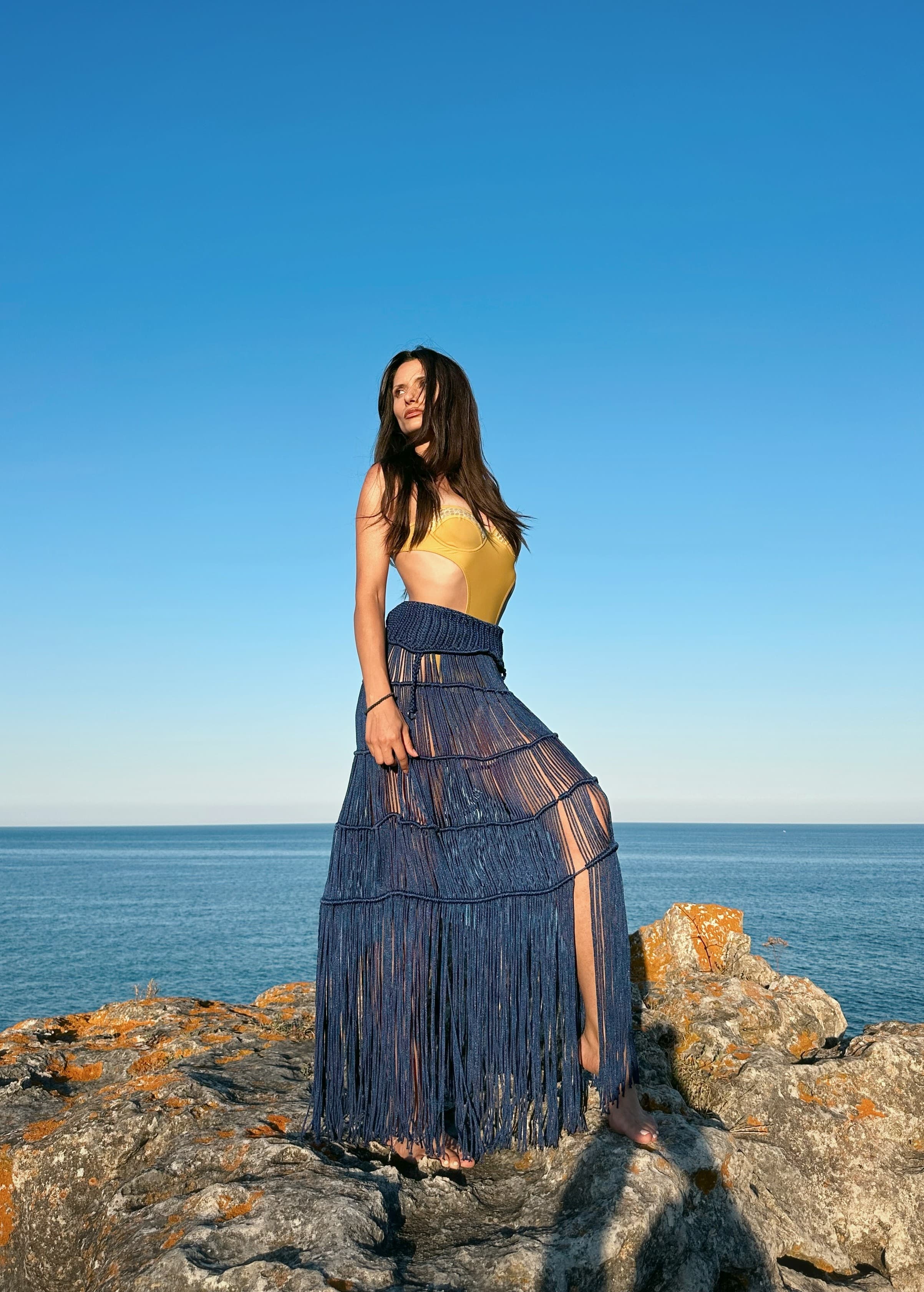 A woman wearing a yellow swimsuit and blue fringed maxi skirt standing on coastal rocks near the sea.