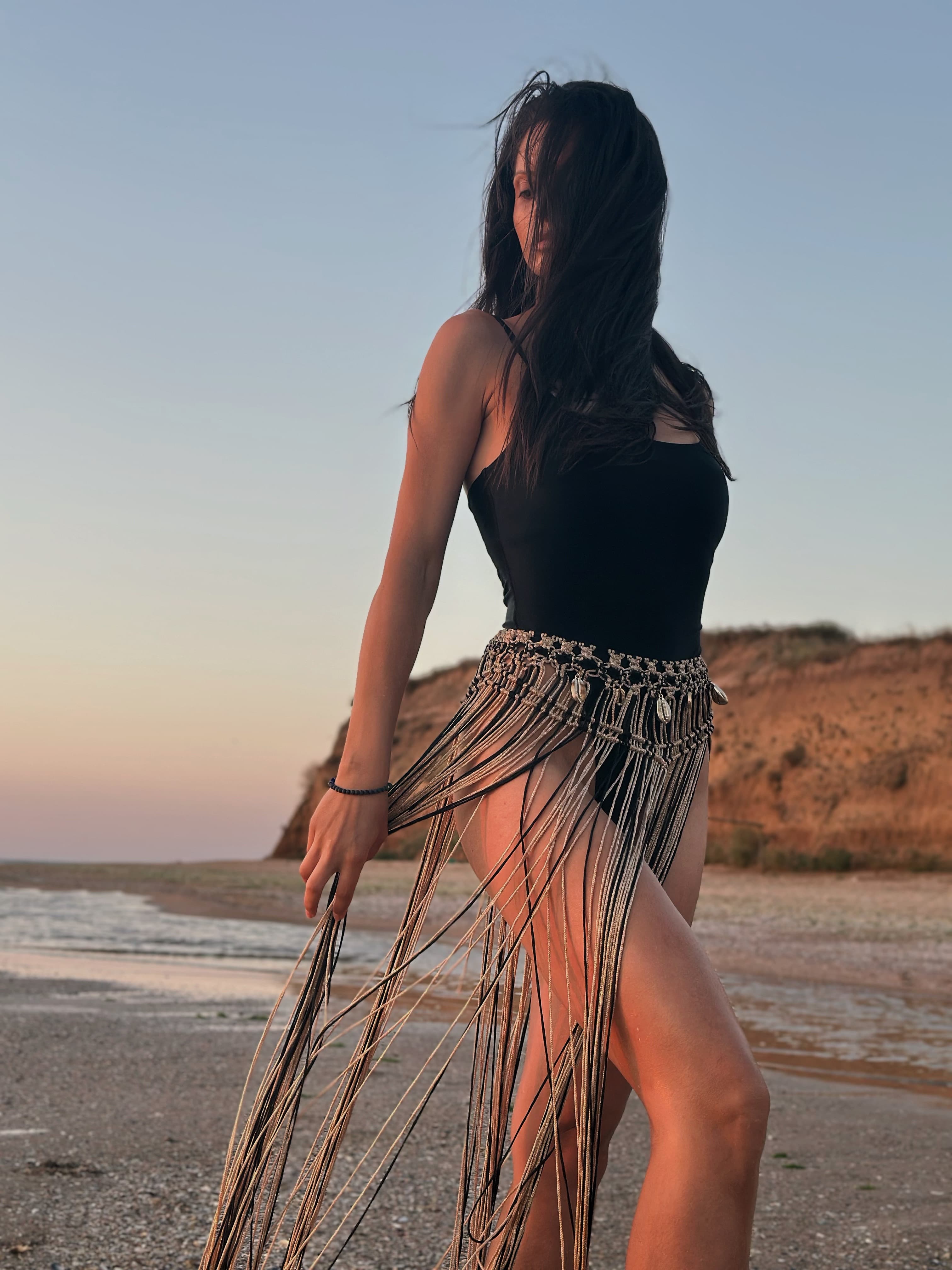 A woman on a beach wearing a black swimsuit and a long gold fringe skirt with shell accents.