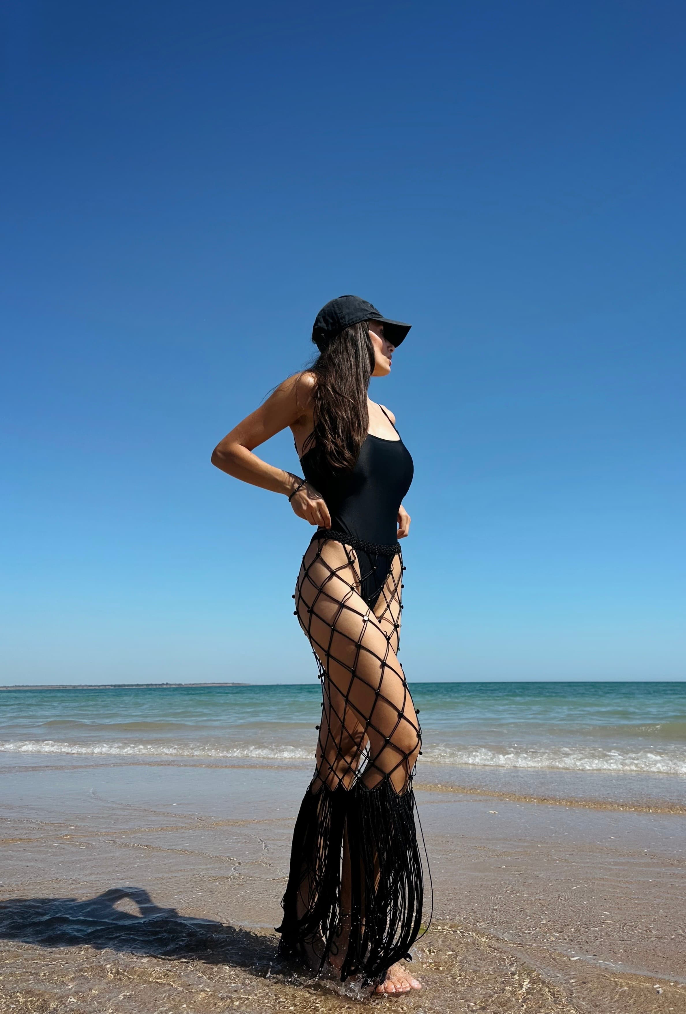 A woman wearing a black swimsuit and layered fringe skirt stands on a beach.