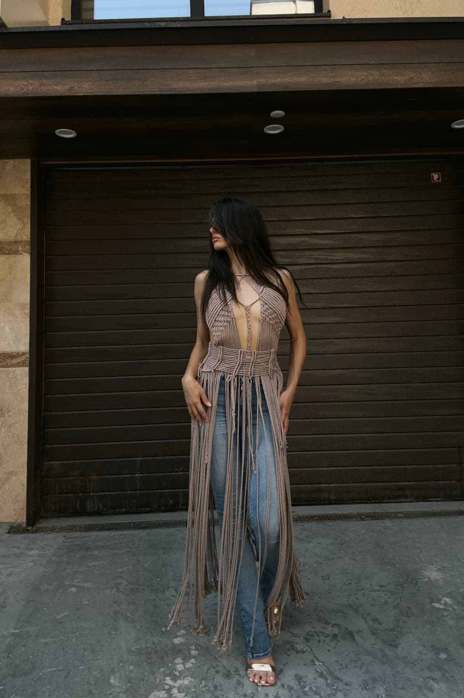 A woman stands wearing a taupe macrame halter top with long fringe over blue jeans.