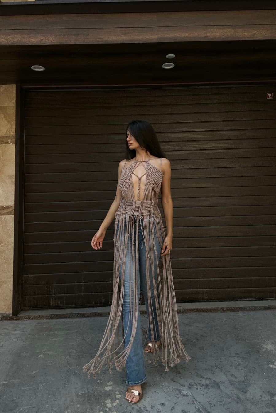 A woman wearing a beige macrame dress over blue jeans stands outside in front of a dark garage door.