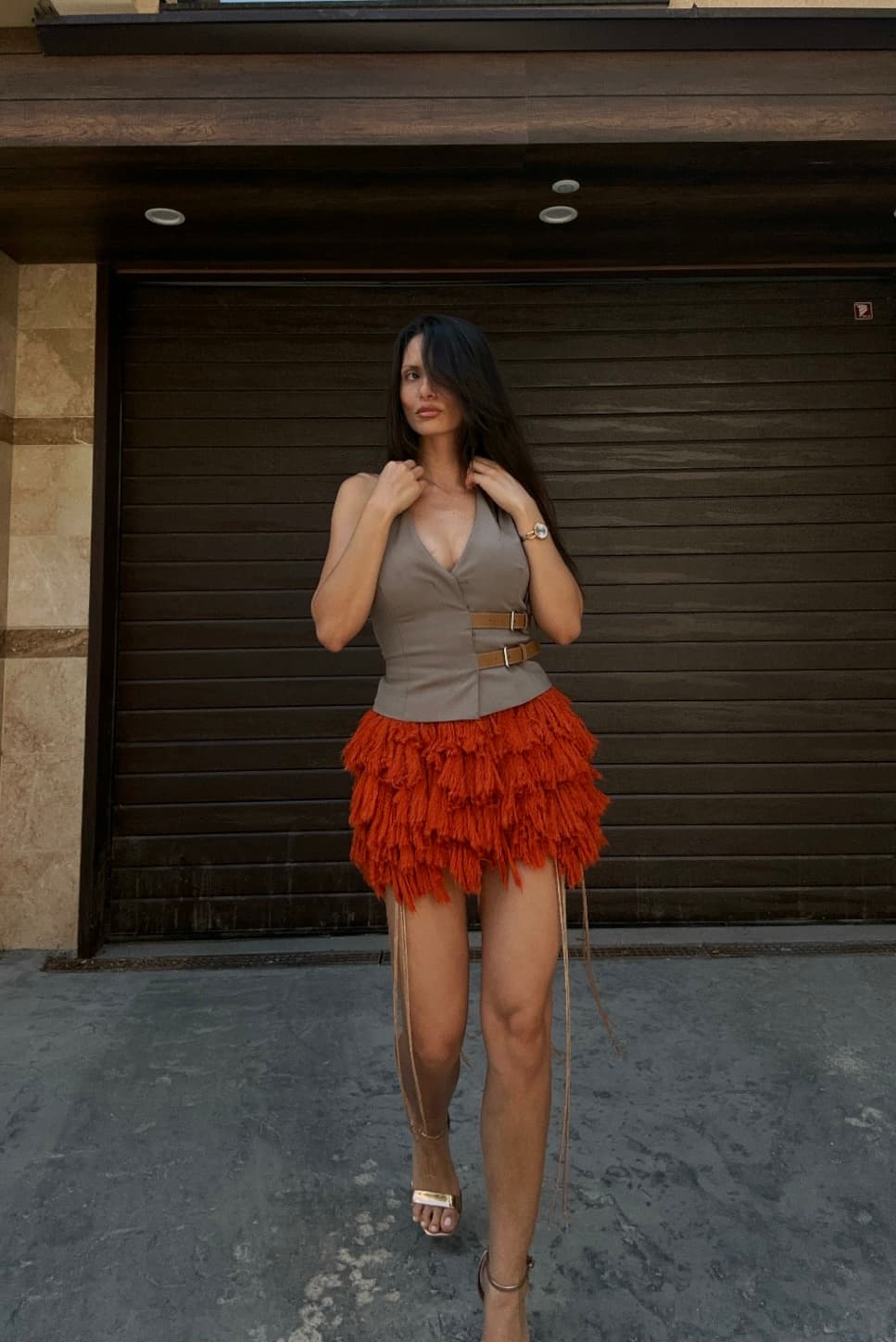 A woman wearing a grey vest and a bright orange tiered fringe mini skirt.