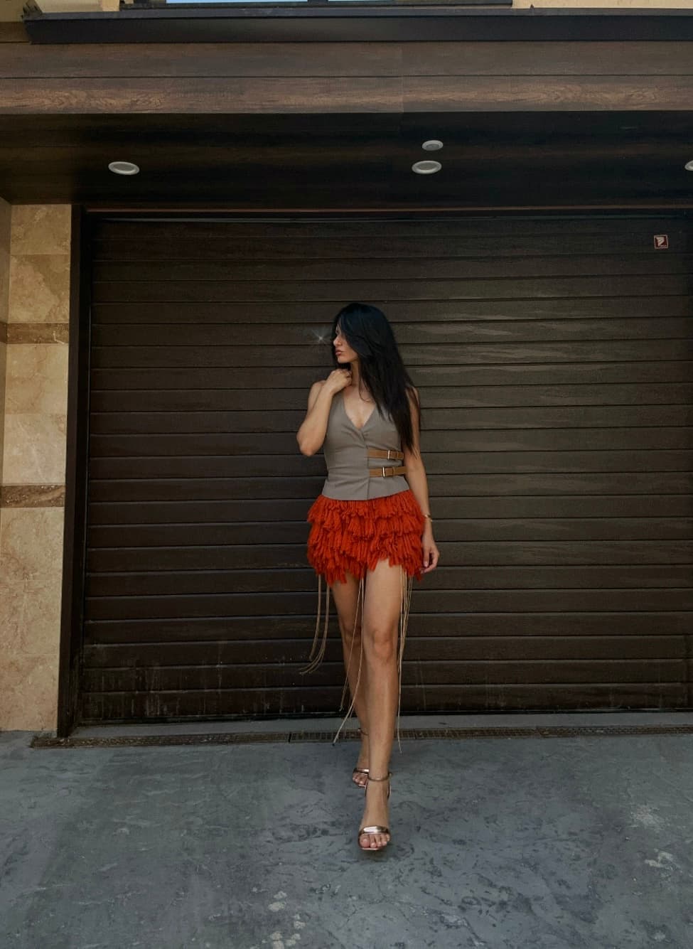 A woman models a brown vest and an orange tiered fringe mini skirt, standing against a dark garage door.