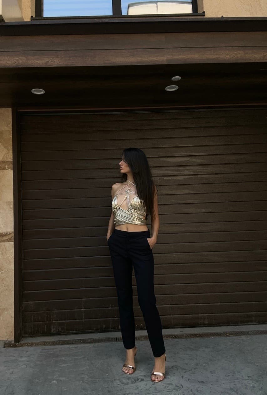 A woman stands in front of a dark garage door, wearing a shiny golden metallic bustier top and dark trousers.